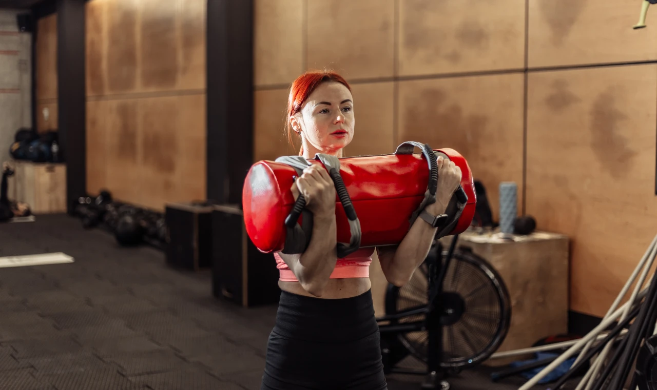 Zercher Carry: Hold a sandbag in the crook of your elbows and perform a Farmers Walk. Woman holding a sandbag in the crook of her elbows while performing a Farmers Carry